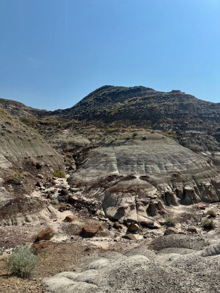 Arid canyon landscape with exposed rock layers, eroded hillsides, sparse vegetation, and a dry streambed under clear blue sky