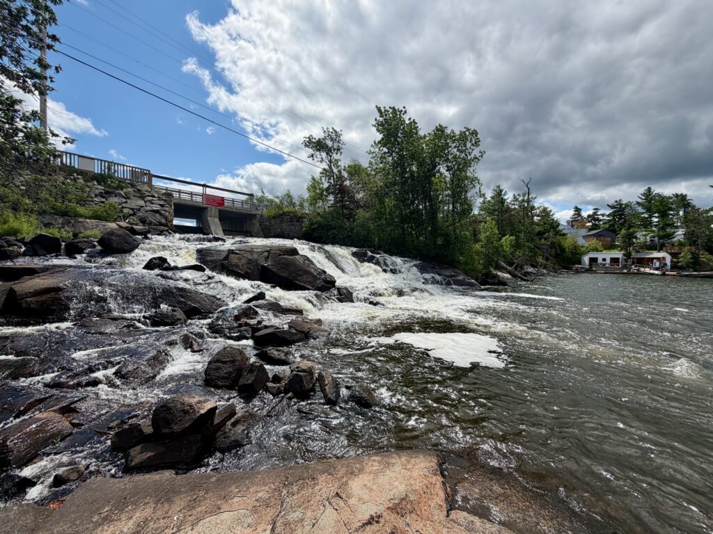 Rushing water flows over rocky rapids beneath a bridge spanning a river, with forested banks and scattered buildings visible along the shoreline under a partly cloudy sky