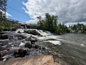 Rushing water flows over rocky rapids beneath a bridge spanning a river, with forested banks and scattered buildings visible along the shoreline under a partly cloudy sky