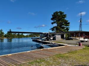 A blue and white floatplane with pontoons is docked at a wooden pier on a calm lake, with a small red hangar and communications tower visible on the shoreline