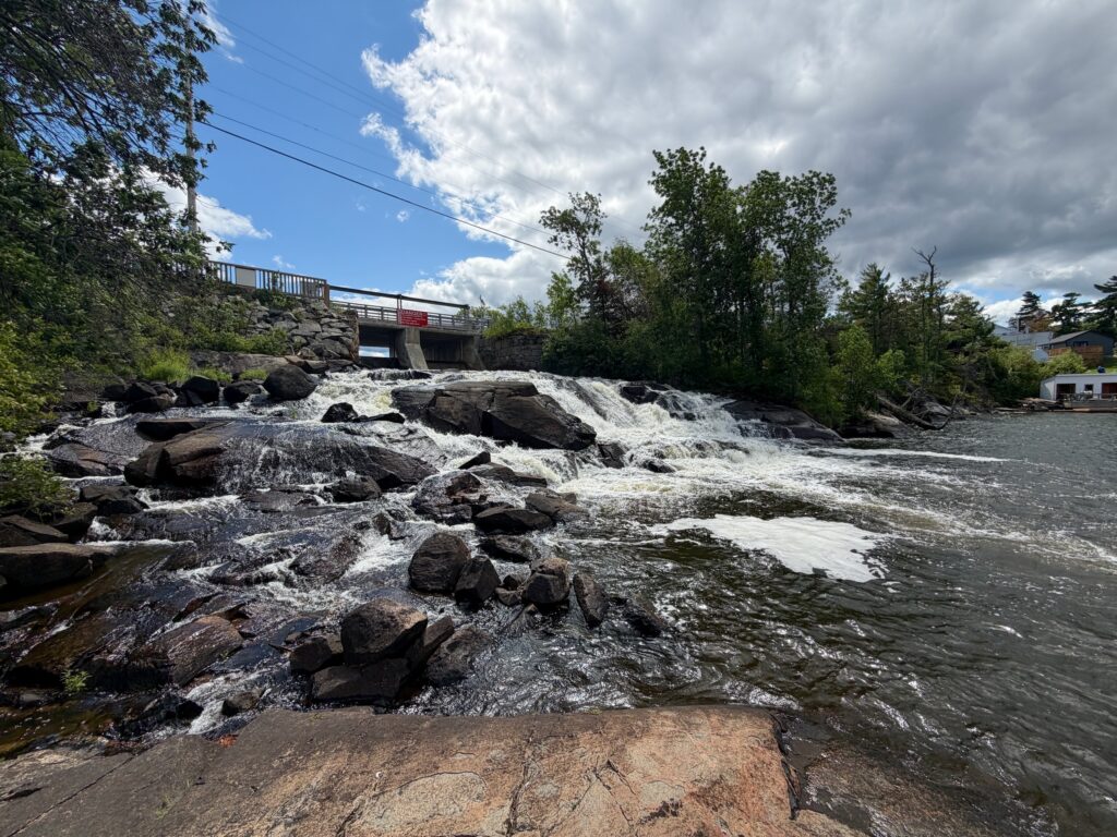 Turbulent river flowing over rocky terrain with a concrete bridge spanning across in the background, surrounded by dense green trees and partly cloudy sky
