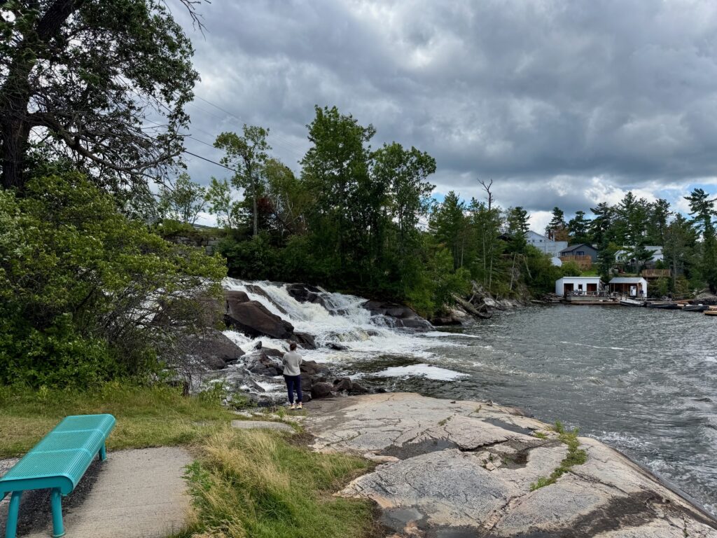 Person standing on rocky shoreline near waterfall with turquoise bench in foreground, cottage visible across the water under cloudy sky