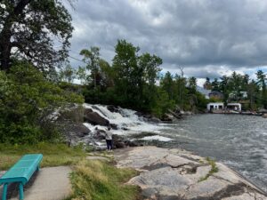 Person standing on rocky shoreline near waterfall with turquoise bench in foreground, cottage visible across the water under cloudy sky