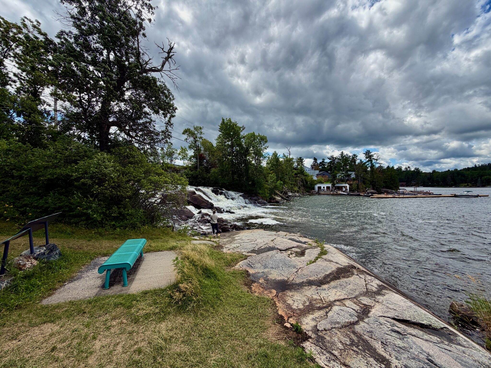Waterfront park with a turquoise bench on a gravel path overlooking a river, rocky shoreline, small waterfall, forested banks, cottages across the water, and stormy sky above