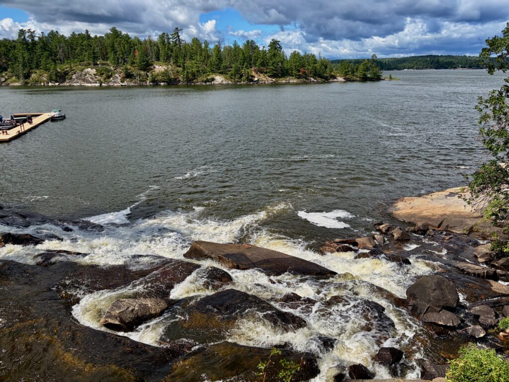 Rocky shoreline with waves breaking against boulders, coniferous forest lining the distant shore, and a moored boat visible to the left under partly cloudy sky