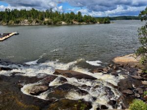 Rocky shoreline with waves breaking against boulders, coniferous forest lining the distant shore, and a moored boat visible to the left under partly cloudy sky