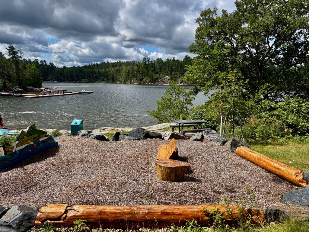 Lakeside beach area with gravel shore, wooden logs, a fire pit, picnic table, and boats on the water surrounded by coniferous forest under partly cloudy sky