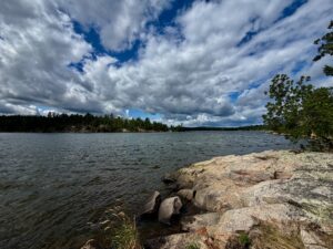 Rocky shoreline with granite boulders beside a calm lake, coniferous forest lining the far shore, and dramatic white clouds against blue sky
