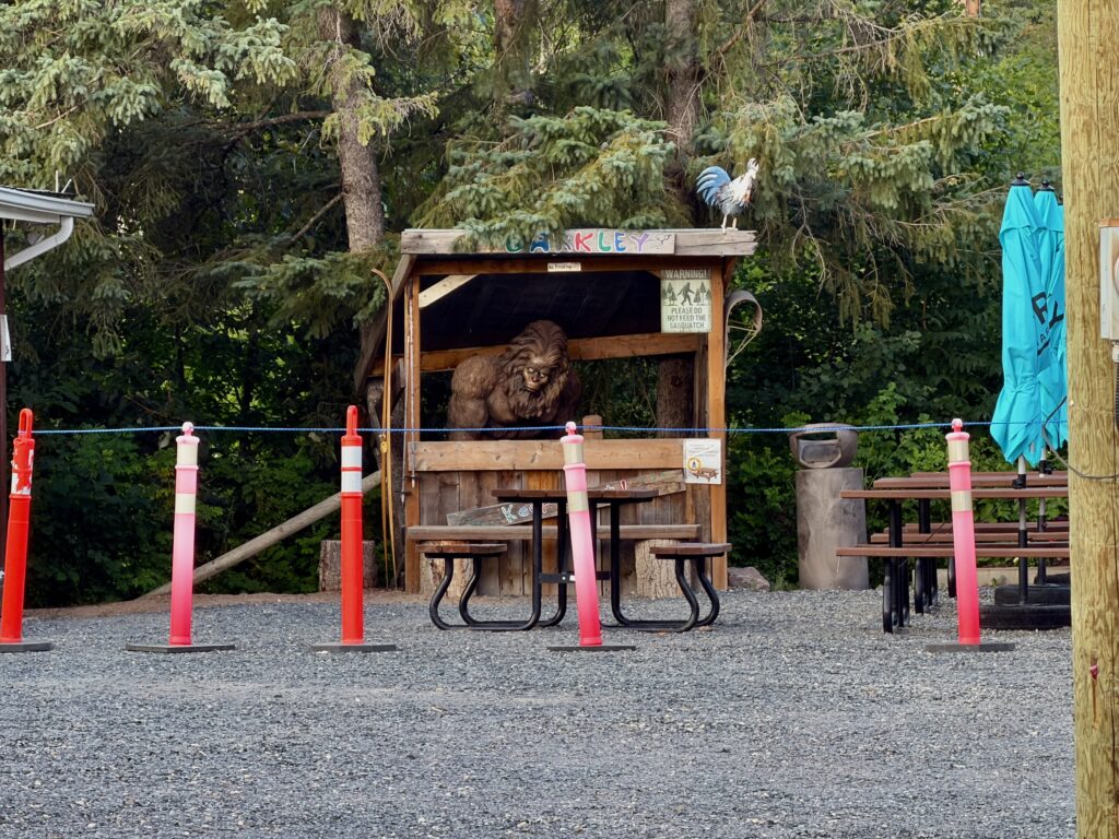 A wooden enclosure with a rooster perched on top and a large brown lion visible inside, surrounded by a red and pink rope barrier in a forested setting
