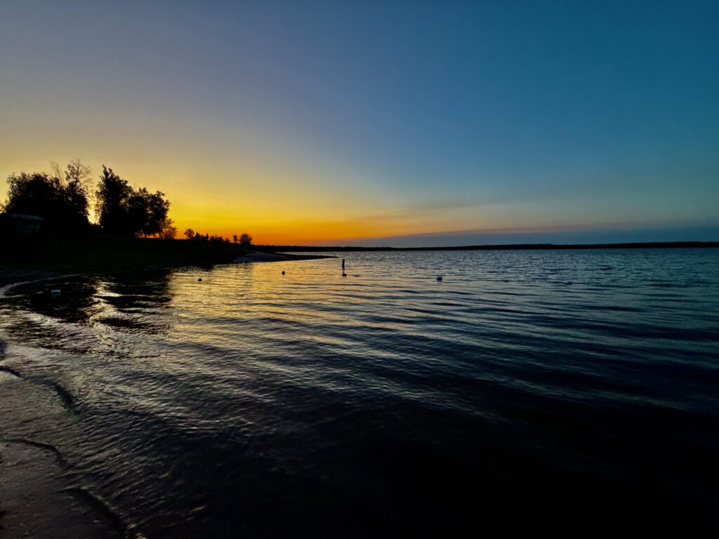 Sunset over a calm lake with silhouetted trees along the shoreline and buoys marking the water