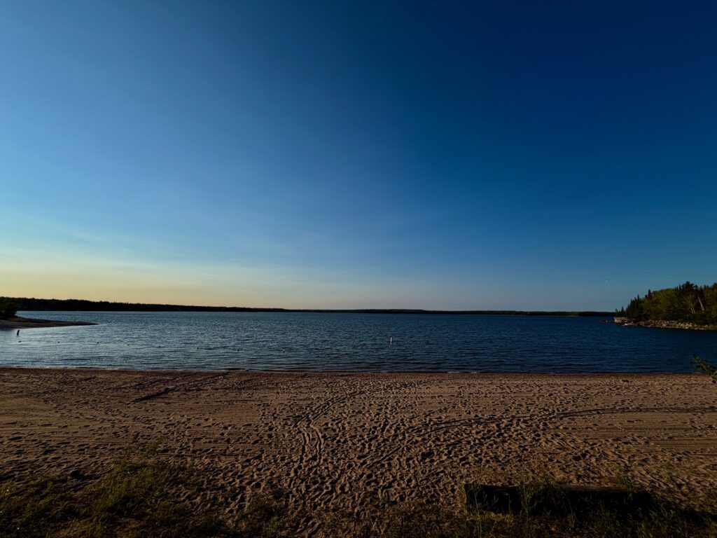 Sandy beach on a calm lake with forested shoreline under clear blue sky