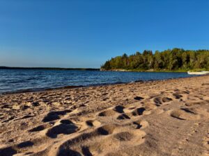 Sandy beach with footprints in the foreground, calm blue water, and a forested shoreline under clear sky