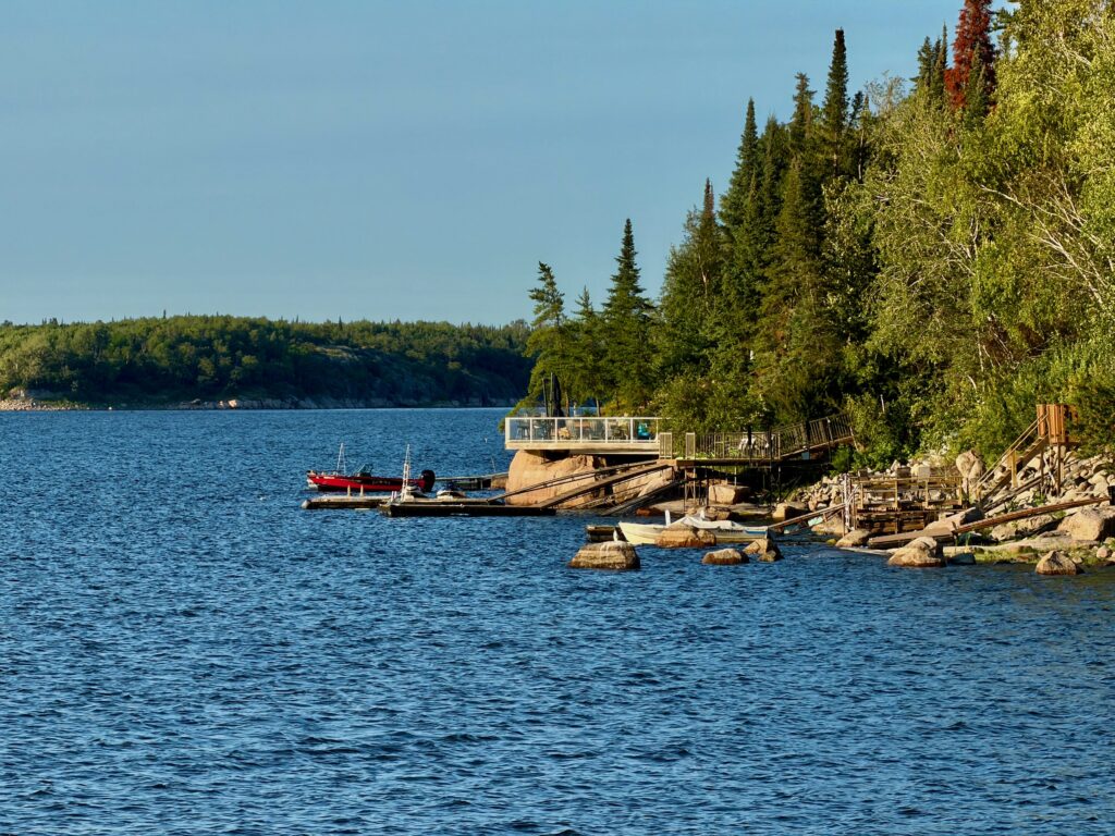 Boathouse and docks on a rocky shoreline with moored boats, surrounded by coniferous and birch trees under clear blue sky
