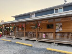 Rustic wooden general store with covered porch, signage for groceries, snacks, clothing and giftware, Canadian flags, and idle-free zone sign in parking area