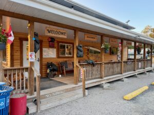 Rustic wooden lodge storefront with "Welcome" sign, carved black bear sculptures on porch, "Open" neon sign, Canadian flag, and signs for Firewood and Smores. Wooden railings and steps lead to entrance marked "No Parking