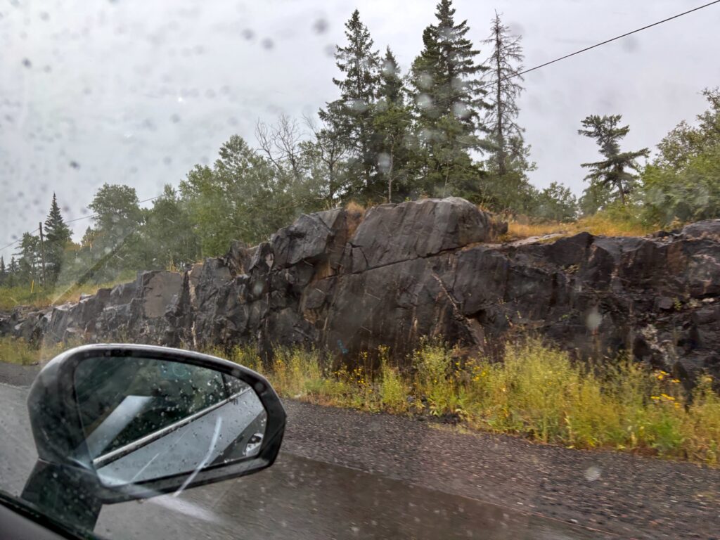 Car mirror reflecting a rainy mountain road with rocky outcrop and coniferous forest in British Columbia