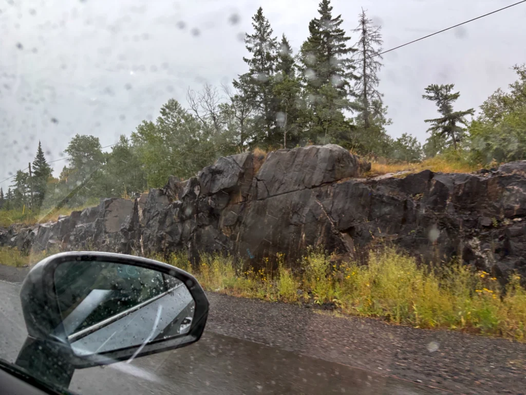 Car mirror reflecting a rainy mountain road with rocky outcrop and coniferous forest in British Columbia