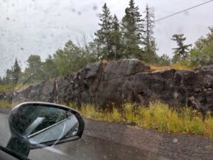 Car mirror reflecting a rainy mountain road with rocky outcrop and coniferous forest in British Columbia