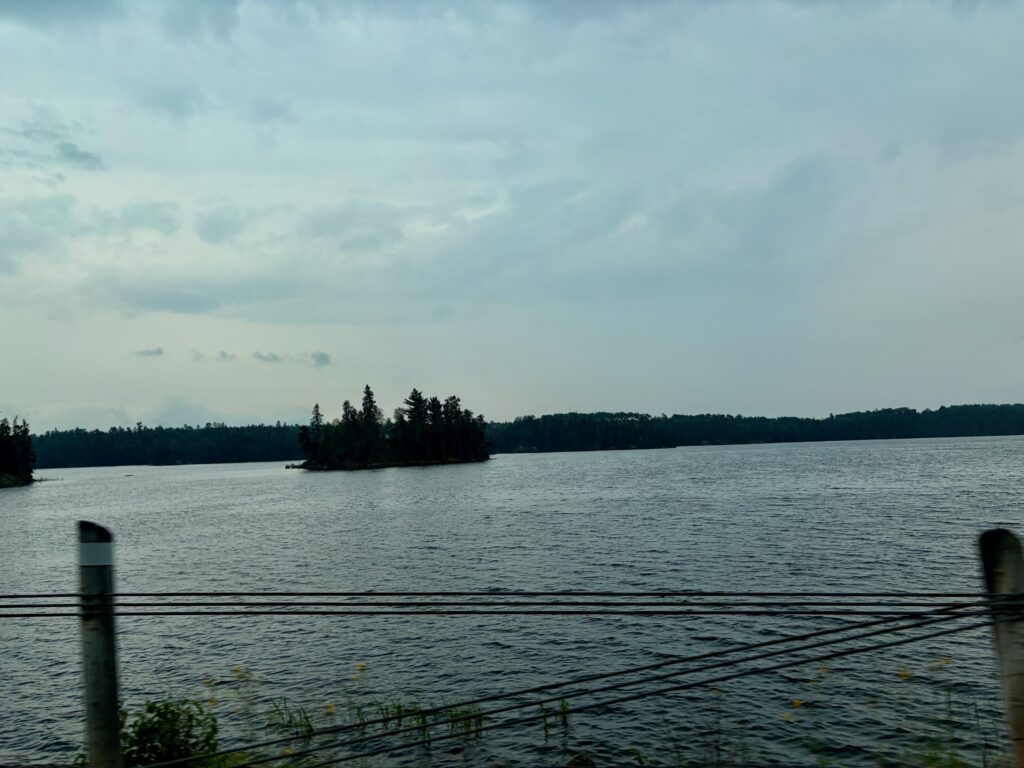 A calm lake with a small forested island in the distance, viewed from a dock with mooring posts, under an overcast sky