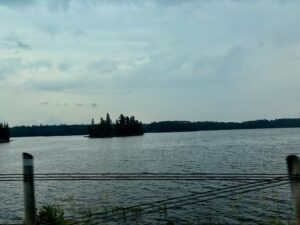 A calm lake with a small forested island in the distance, viewed from a dock with mooring posts, under an overcast sky