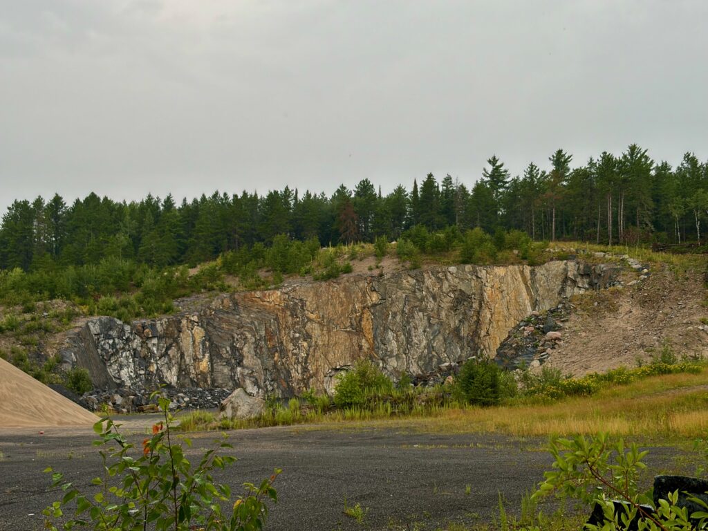 Abandoned quarry with exposed rock face and layered stone formations surrounded by coniferous forest and vegetation under overcast sky