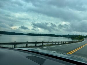 View from a vehicle windshield of a highway curving along a calm lake under overcast skies, with coniferous forest lining the far shore
