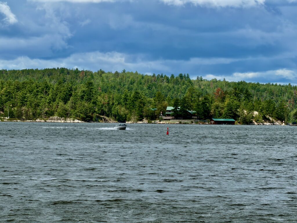 Motorboat on a lake with coniferous forest and cottages lining the rocky shoreline under a partly cloudy sky