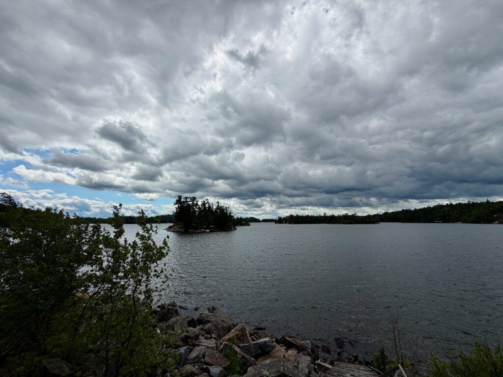 Lake surrounded by coniferous forest and rocky shoreline under dramatic cloud-filled sky with patches of blue