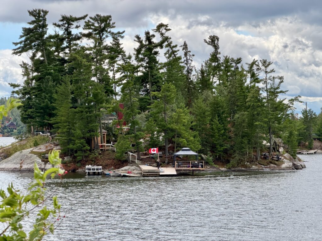 Waterfront cottage nestled among tall pine trees with a Canadian flag on the dock, situated on a rocky island in a Canadian lake under partly cloudy skies