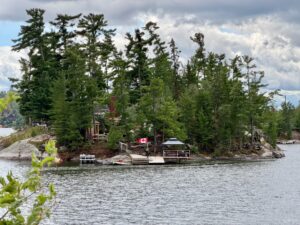 Waterfront cottage nestled among tall pine trees with a Canadian flag on the dock, situated on a rocky island in a Canadian lake under partly cloudy skies