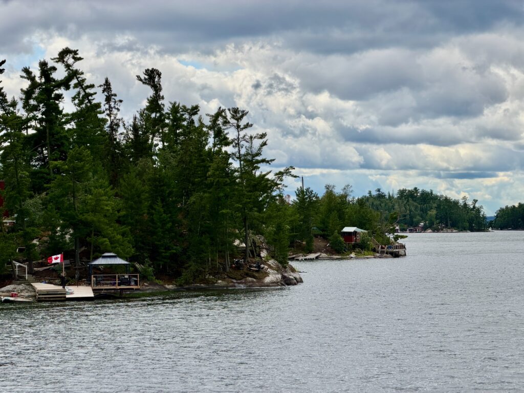 Waterfront cottages nestled among coniferous trees on a rocky shoreline, with a Canadian flag visible on the left cabin and forested islands extending across the lake under a partly cloudy sky