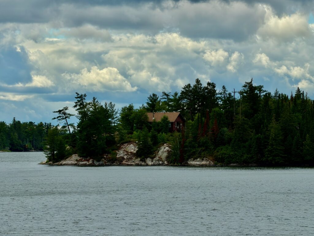 Red brick house with tan roof on a forested rocky shoreline, viewed across calm water under a cloudy sky