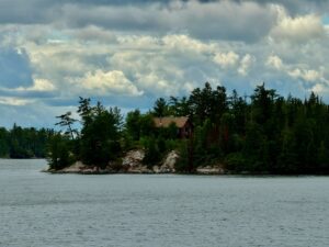 Red brick house with tan roof on a forested rocky shoreline, viewed across calm water under a cloudy sky