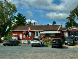 Roadside restaurant with red-and-white striped awning, Canadian and Ukrainian flags, parked vehicles in front, surrounded by trees and blue sky with clouds