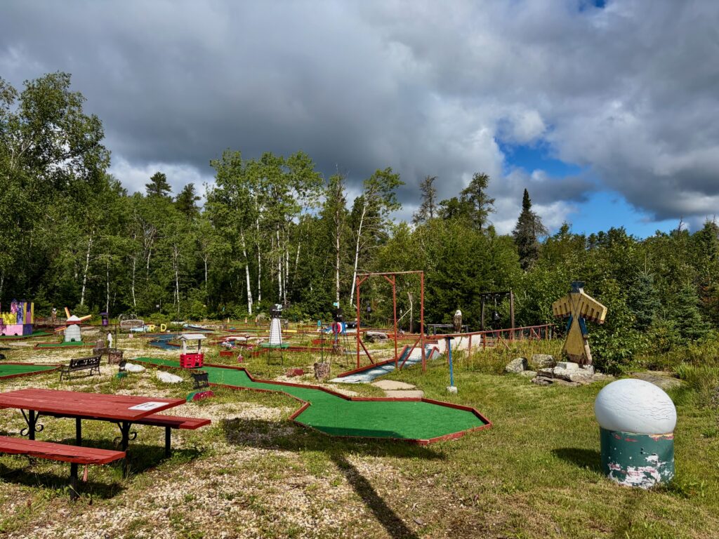 Outdoor mini-golf course with green fairways, red picnic tables, red metal swing set, and painted obstacles, surrounded by boreal forest and cloudy sky