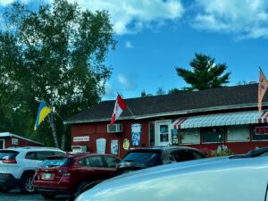 Red restaurant building with Canadian and Ukrainian flags, parked cars in front, and tall trees under blue sky