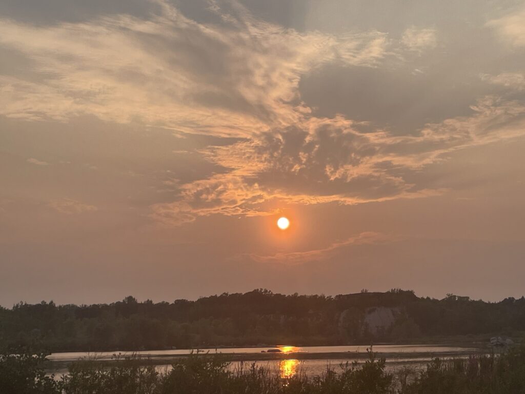 Sunset over a calm lake with forested shoreline, golden sun reflecting on the water beneath a sky of orange and grey clouds