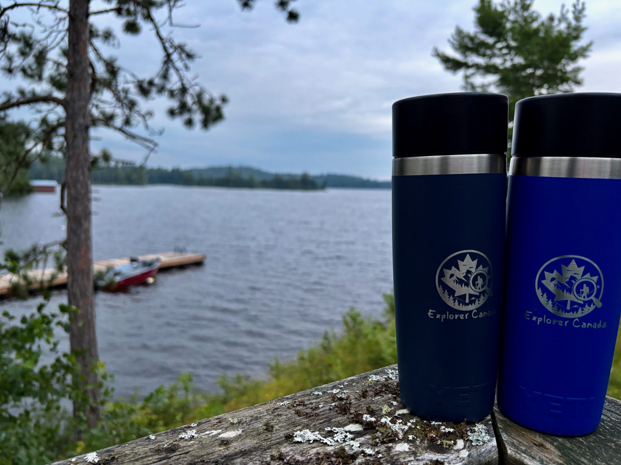Two Explorer Canada branded insulated water bottles sitting on a log overlooking a lake with coniferous trees and a canoe in the background