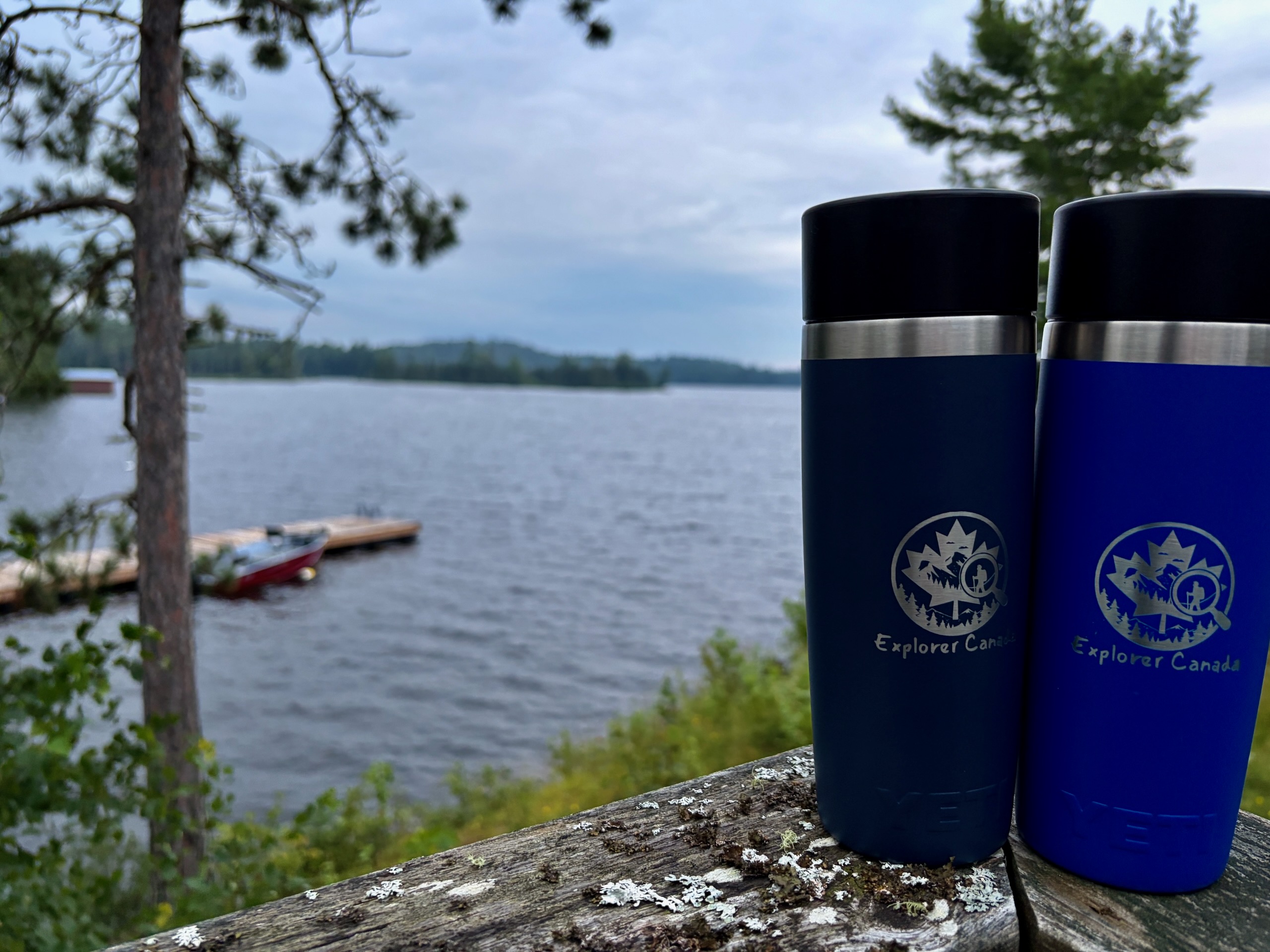 Two Explorer Canada branded insulated water bottles sitting on a log overlooking a lake with coniferous trees and a canoe in the background