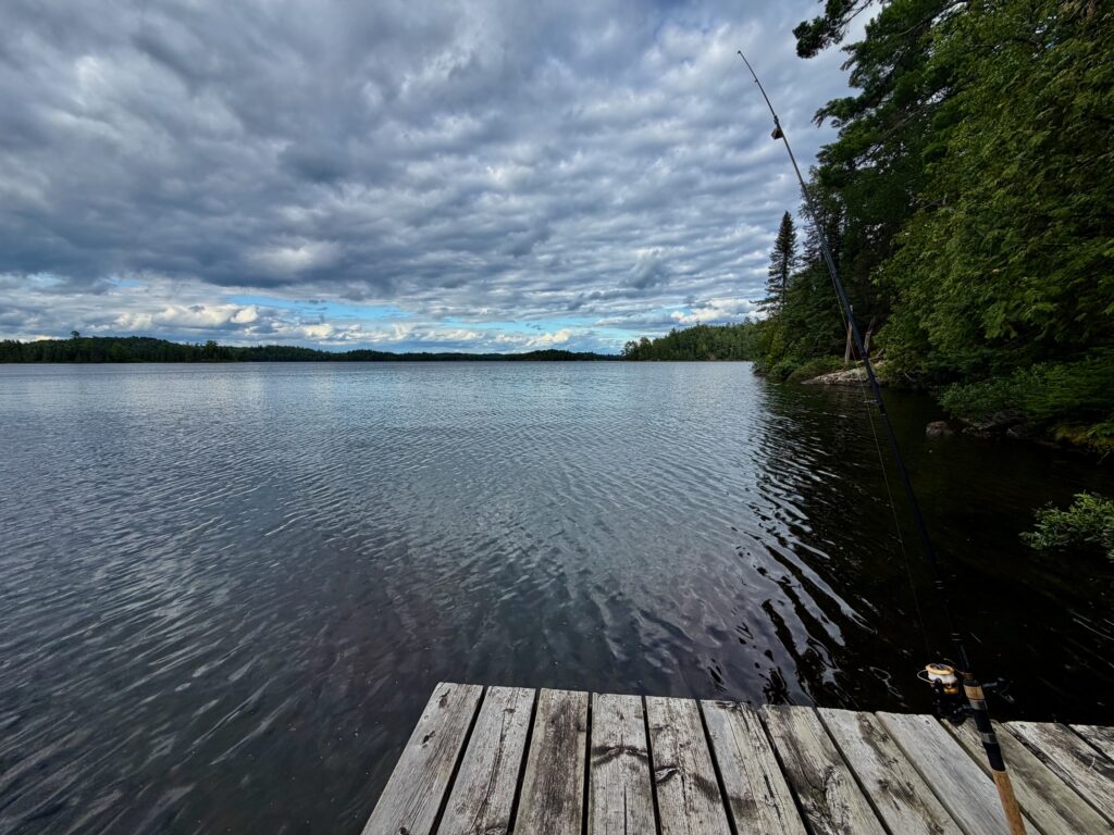Wooden dock extending into a calm lake with a fishing rod set up, surrounded by dense forest and cloudy sky