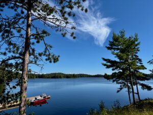 A red and black boat moored on a sandy beach beside a calm blue lake, framed by tall pine trees under a clear sky with wispy clouds