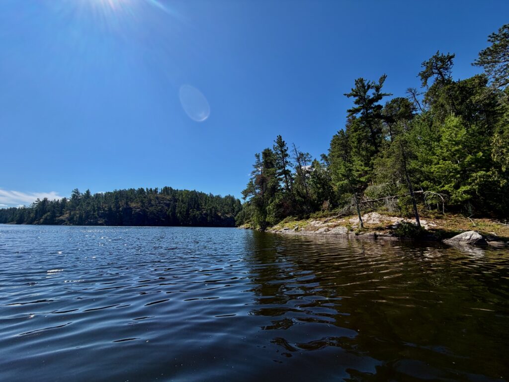 A pristine lake with clear blue water reflecting the sky, surrounded by dense coniferous forest and rocky shoreline under bright sunshine