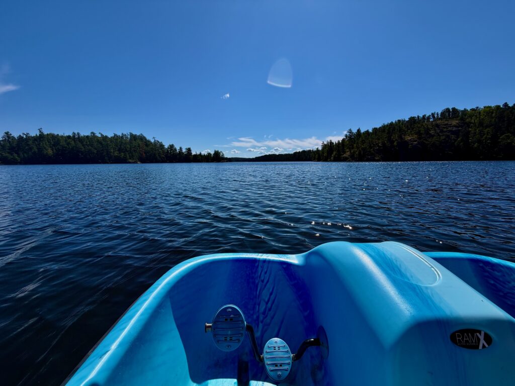 View from inside a blue kayak on a calm lake surrounded by coniferous forest under clear sky