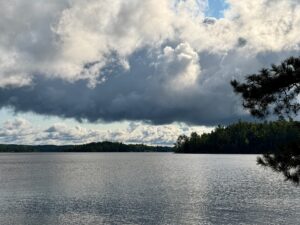 Lake surrounded by forested shoreline beneath a dramatic sky with billowing white and grey clouds, with pine branches visible on the right edge