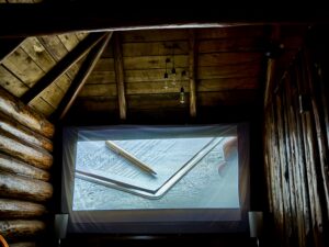 Projector screen displaying a close-up of architectural blueprints and pencils on a textured surface, mounted in a rustic wooden cabin interior with exposed beams and hanging Edison bulbs