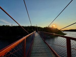 Suspension bridge with red railings and mesh fencing spanning a river at sunset, forested shoreline visible on both sides