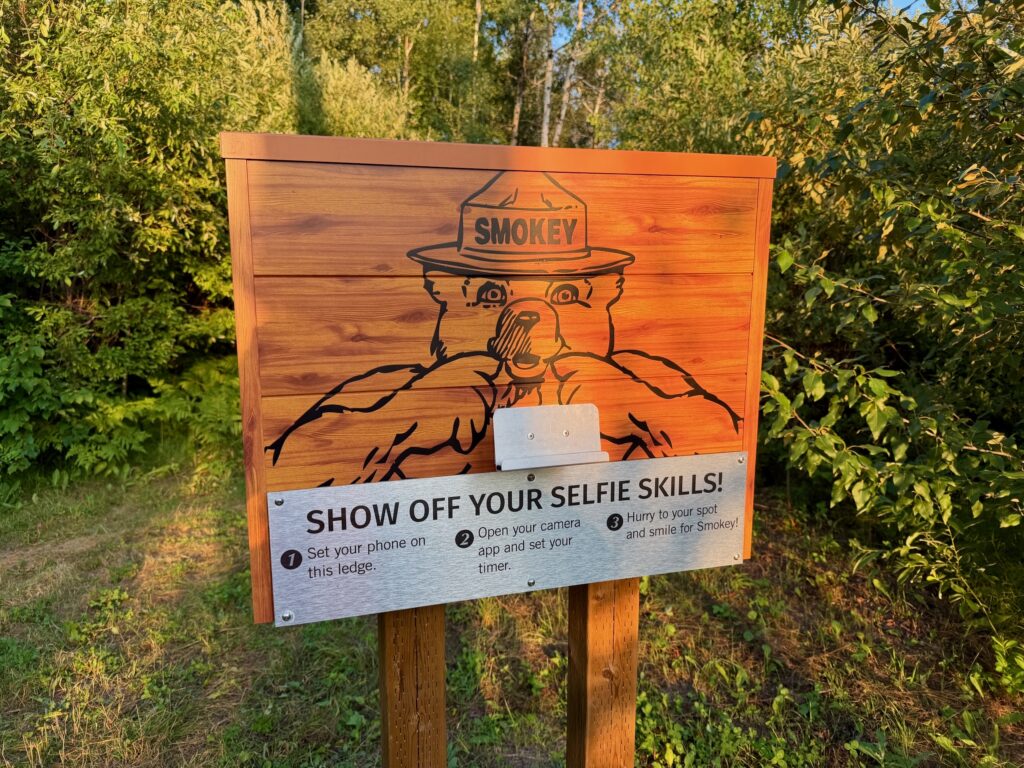 Wooden sign featuring Smokey the Bear wearing a ranger hat, with instructions to take a selfie: set phone on ledge, open camera app and set timer, then smile for Smokey
