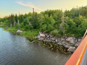 Rocky shoreline with boreal forest of spruce, birch, and pine trees along a calm lake, viewed from a boat at sunset
