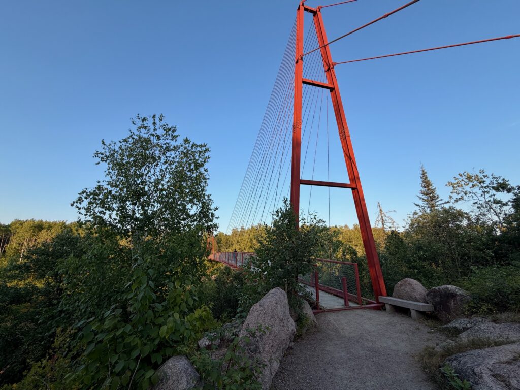 Red cable-stayed bridge with tall pylon spanning forested landscape under clear blue sky, large boulders in foreground