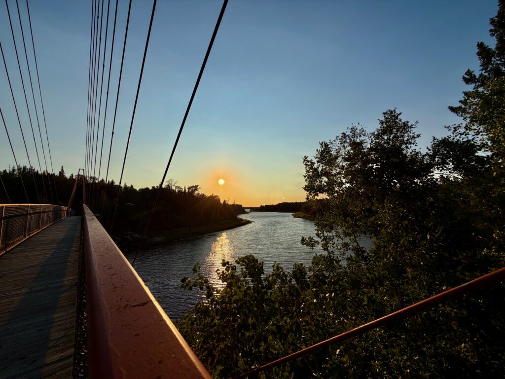 Suspension bridge walkway at sunset overlooking a river with forested shorelines and golden light reflecting on the water
